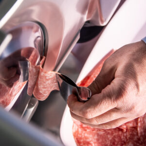 Mano con pinza prende una fetta di salame che esce dalla lama di un'affettatrice.